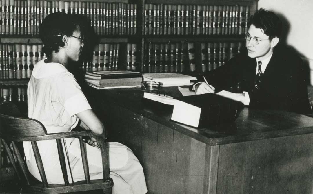 Black and white photo of man behind a desk speaking to a woman.