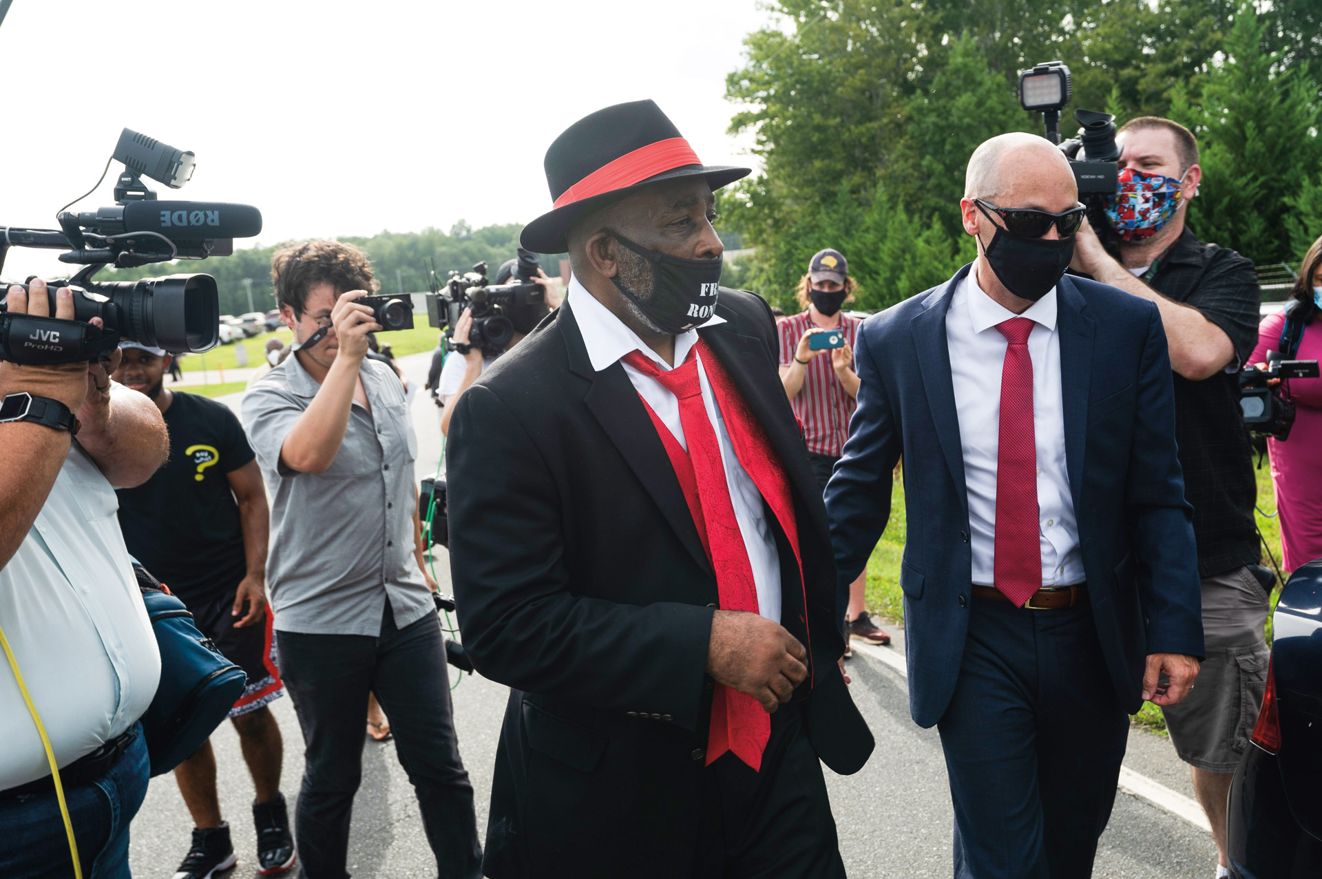 Man in suit with red tie and red band around hat walking past TV cameras